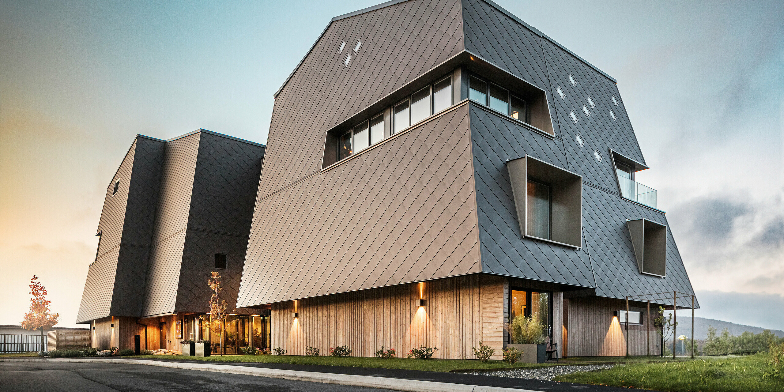 Vue de l'entrée de l'hôtel Beckerwyc à Bekkjarvik, en Norvège, au crépuscule. La façade est revêtue de losanges PREFA 44 × 44 en brun noisette, ce qui confère au bâtiment un aspect à la fois moderne et impressionnant. L'éclairage met en valeur les détails architecturaux et la forme inhabituelle du bâtiment. La combinaison d'un design futuriste et de couleurs naturelles s'intègre harmonieusement dans l'environnement, souligné encore par les conditions d'éclairage agréables.