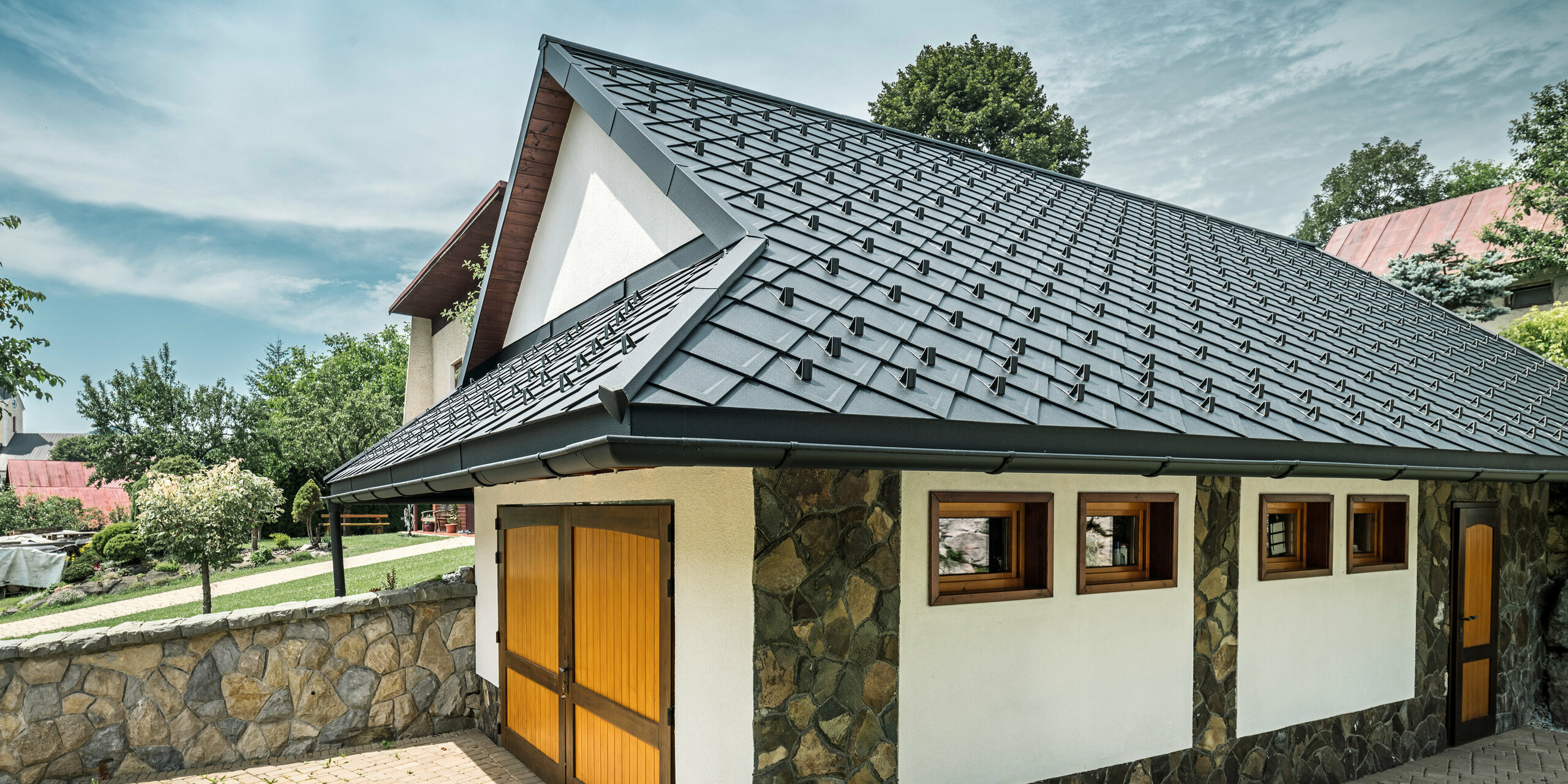 Garage d'une maison familiale traditionnelle à Stara Bystrica, Slovaquie, avec un toit en losanges PREFA 29 × 29 en P.10 noir. La couverture de toiture robuste en aluminium s'harmonise parfaitement avec la façade en pierre naturelle du garage. Une allée pavée est visible au premier plan, ce qui souligne l'aspect soigné de l'ensemble de la propriété.