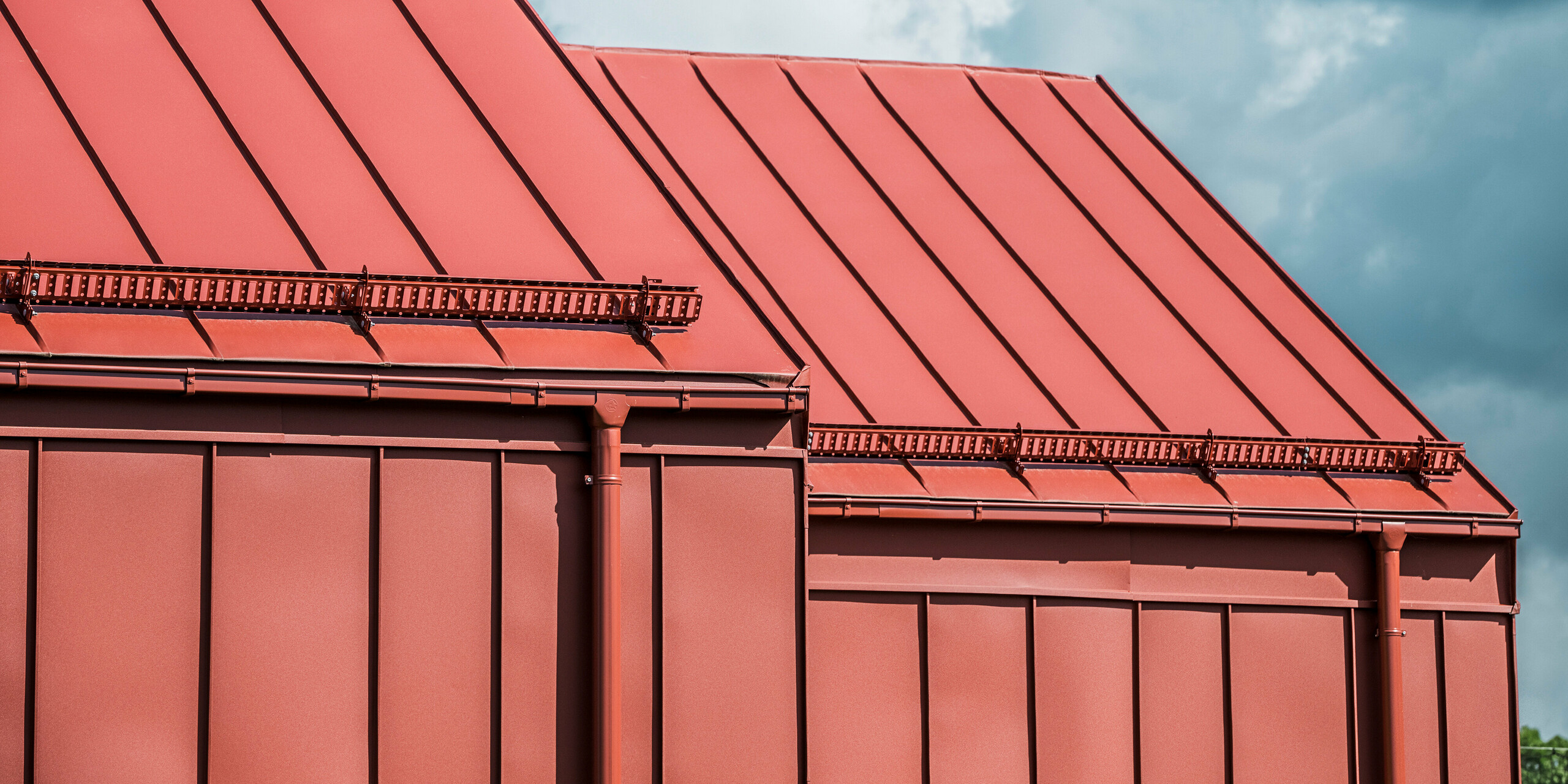 Gros plan sur les avant-toits de la cantine scolaire de Broby, en Suède. Vous pouvez constater le revêtement propre et précis des surfaces de toiture avec PREFALZ en rouge oxyde. Les systèmes d'évacuation des eaux de toiture, y compris la gouttière et le tuyau d'évacuation de PREFA, sont également peints en rouge oxyde et s'intègrent parfaitement dans le design global. Les marches sur le toit offrent une sécurité supplémentaire. Les lignes claires de la toiture à joints debout et la palette de couleurs uniforme soulignent la qualité de fabrication et le design moderne du bâtiment.