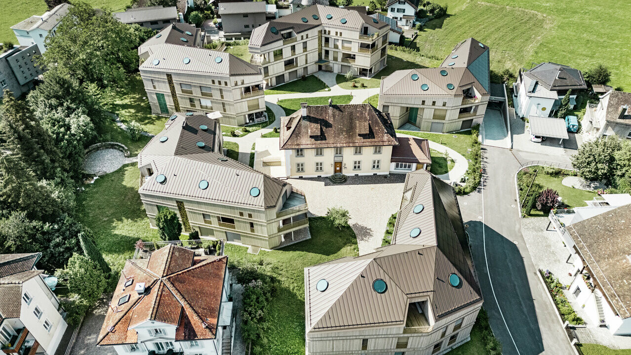 Vue aérienne du complexe résidentiel moderne de Liebwylen, en Suisse. Les bâtiments sont conçus avec des toits en aluminium PREFALZ de couleur brune et disposent de fenêtres de toit rondes. Au centre se trouve la villa historique, entourée de nouveaux bâtiments harmonieusement disposés, le tout intégré dans un paysage verdoyant.