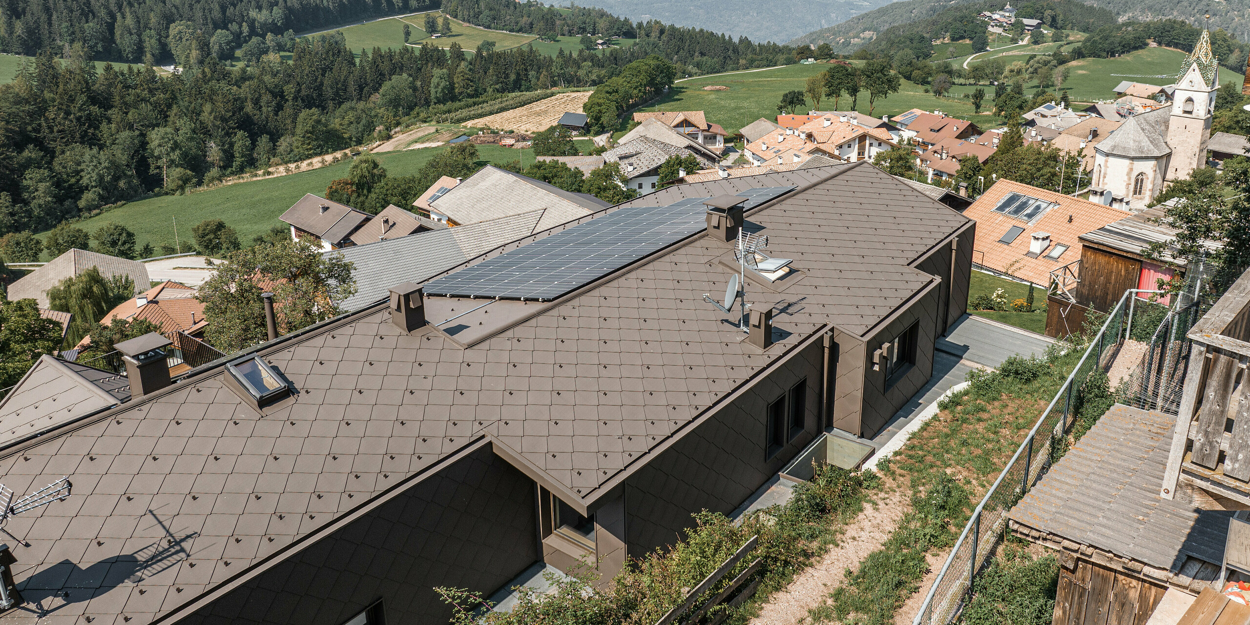 Luftaufnahme eines modernisierten Doppelhauses in Mölten, Südtirol, mit einem Dach aus braunen PREFA Aluminiumrauten. Auf dem Dach wurde auch eine Photovoltaikanlage installiert. Das Haus fügt sich nahtlos in die dörfliche Umgebung mit traditionellen Gebäuden und der Kirche vor der Kulisse eines weiten Tales und grüner Berge ein. Die langlebigen Metallrauten bieten sowohl funktionalen Schutz als auch eine ansprechende Ästhetik im Einklang mit der Natur.