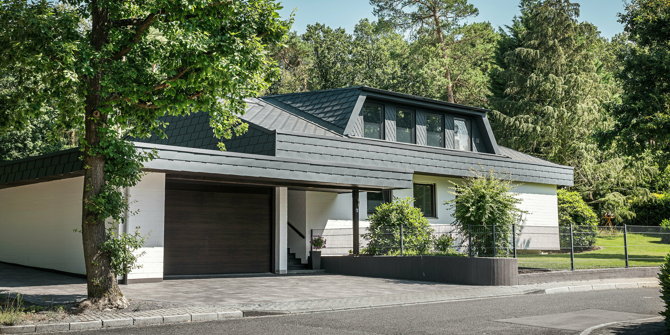 Vue de face d'une maison individuelle à Mechernich avec lucarnes trapézoïdales, habillée de petits losanges de toiture PREFA en P.10 anthracite. Le toit et la zone au-dessus du garage sont recouverts de DS.19 PREFA. La façade blanche de la maison et la porte de garage marron contrastent élégamment avec les éléments de toiture en aluminium sombre. Le jardin de devant est entouré d'arbres et d'arbustes verts, tandis qu'une double clôture en treillis métallique délimite le bâtiment de l'extérieur.