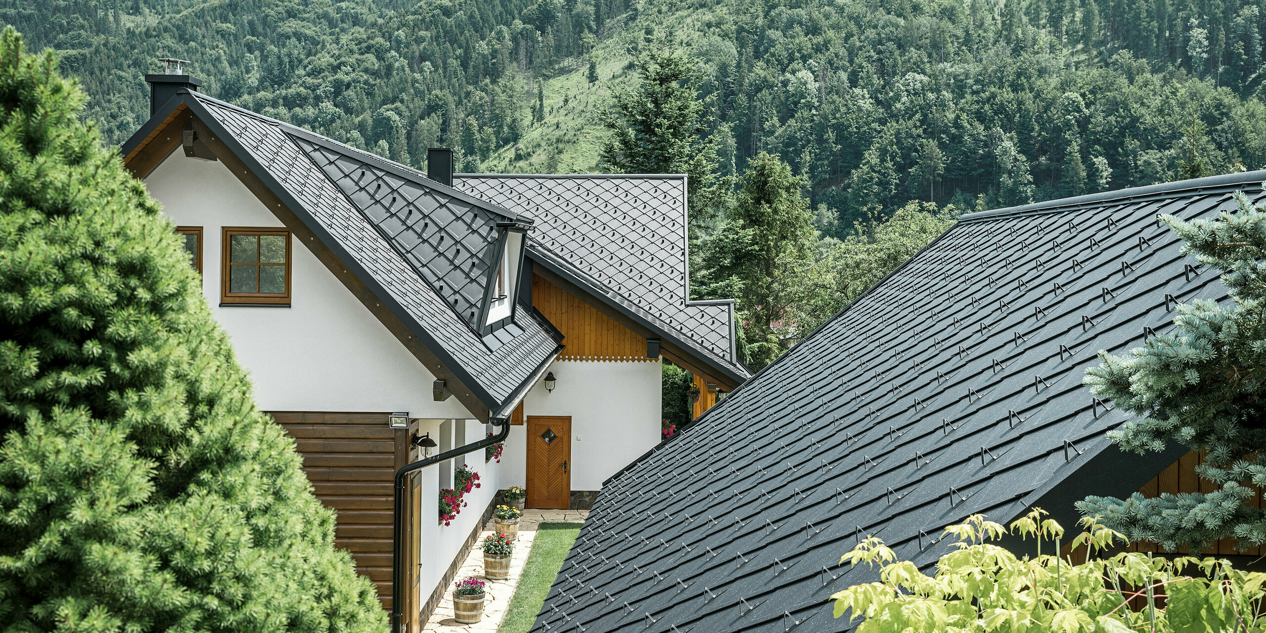 Vue arrière d'une maison familiale traditionnelle à Stara Bystrica, en Slovaquie, avec un paysage de toit à couper le souffle composé de losanges de toit PREFA 29 × 29 en P.10 noir. Les tuiles robustes en aluminium ont un aspect harmonieux avec l'environnement naturel et offrent une modernité contrastée avec un beau parquet au plan. Les surfaces de toit détaillées et les lucarnes trapézoïdales bien intégrées confèrent au bâtiment un aspect esthétique unique.