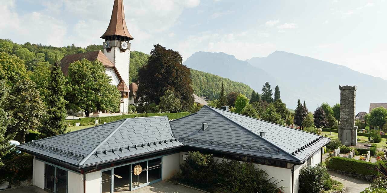 Chambre funéraire avec des panneaux de toiture FX.12 de PREFA couleur anthracite avec, à l’arrière-plan, une église et à côté, le cimetière.