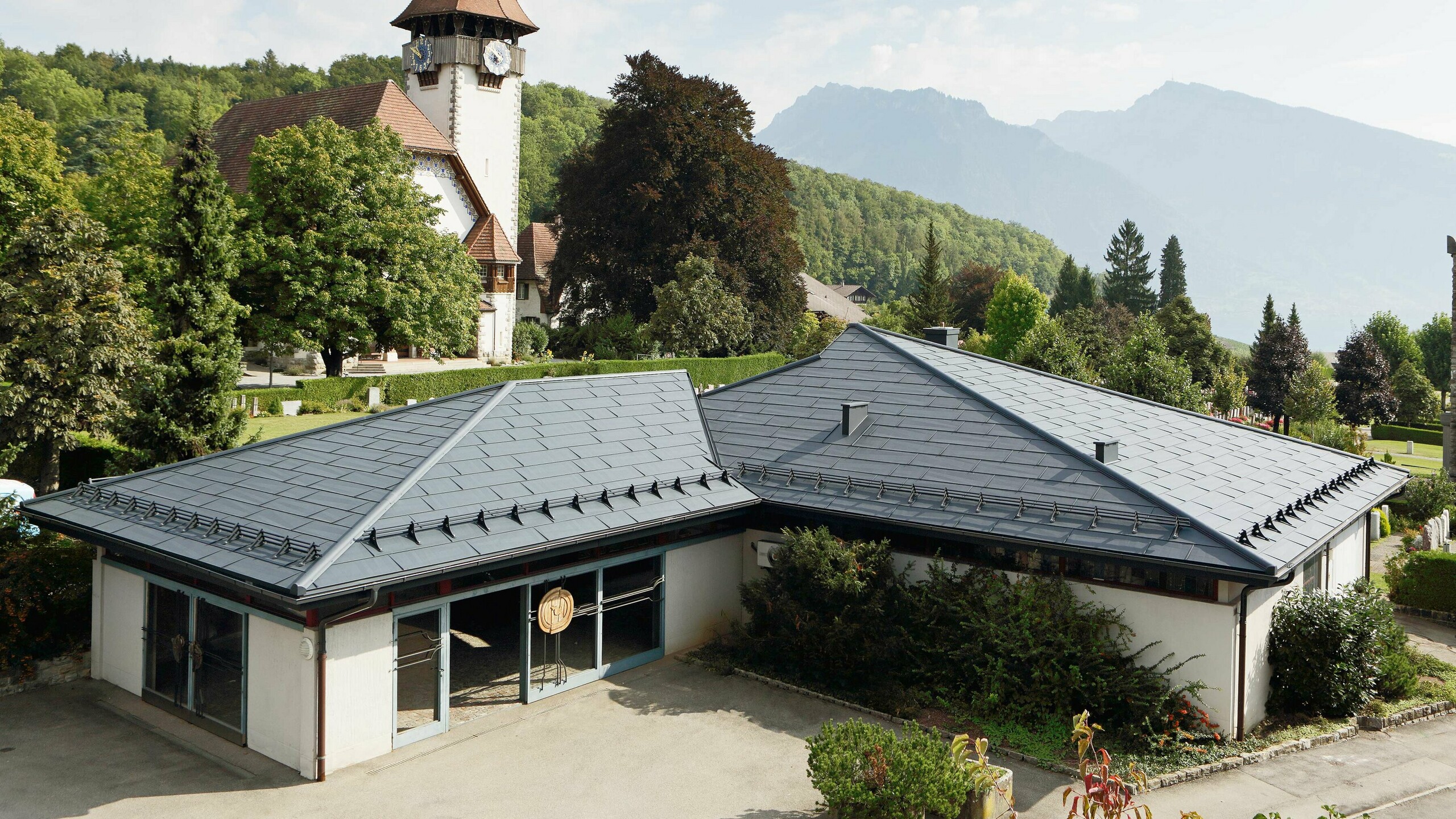 Aufbahrungshalle mit FX.12 Dachpaneelen von PREFA in Anthrazit, im Hintergrund steht eine Kirche und daneben befindet sich der Friedhof.