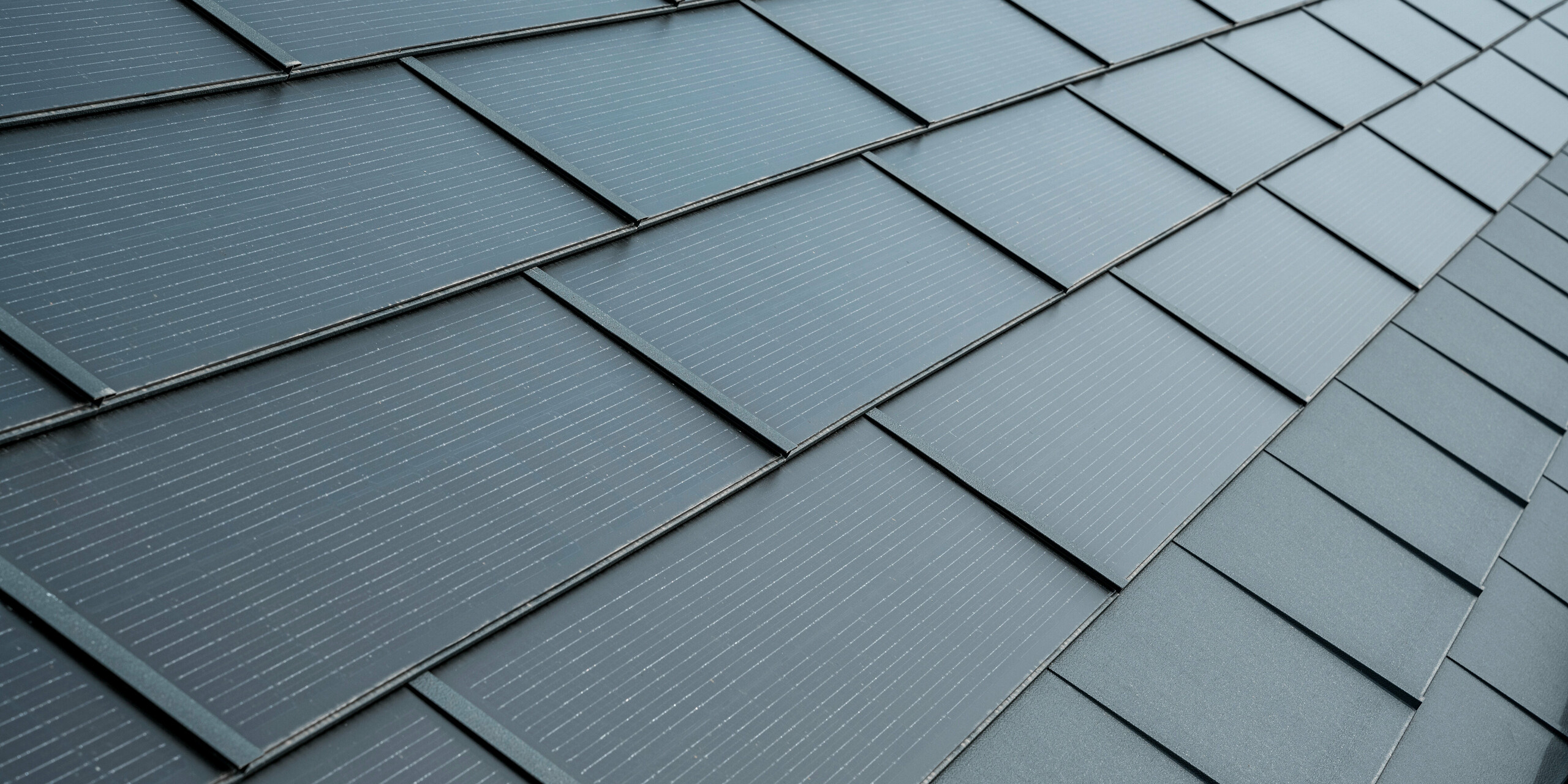 Gros plan sur les tuiles solaires PREFA en anthracite. Les modules solaires sont élégamment intégrés dans le toit et s'intègrent ainsi harmonieusement dans l'ensemble. Les tuiles présentent une surface texturée avec des lignes fines qui apportent une esthétique supplémentaire tout en constituant une solution fonctionnelle et économe en énergie.