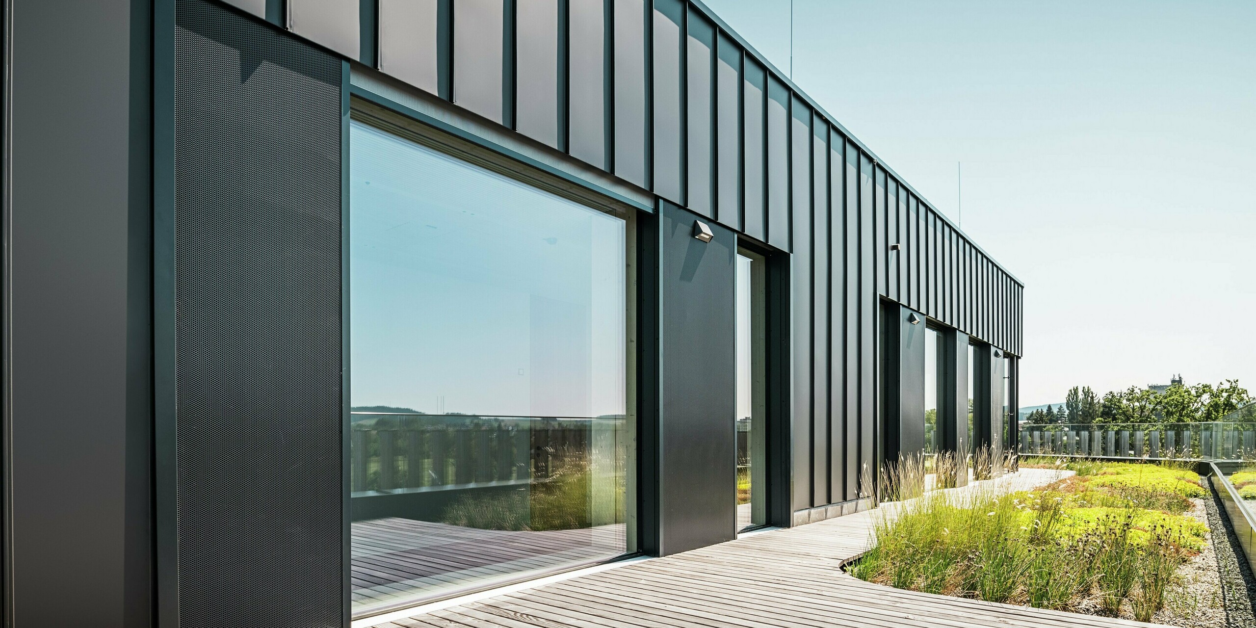 La terrasse sur le toit du Team 7 World à Ried im Innkreis combine une façade PREFALZ en noir-brun avec de grandes façades vitrées et un environnement verdoyant. Le revêtement en aluminium de haute qualité offre durabilité et style moderne, tandis que les éléments naturels du toit végétal assurent un design harmonieux et durable. Idéal pour une architecture contemporaine avec une orientation écologique.