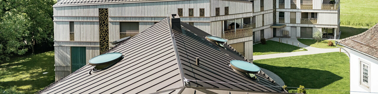 Complexe résidentiel moderne à Liebwylen, en Suisse, avec un paysage de toit innovant en aluminium PREFALZ de couleur brune. Le toit à joint debout intègre de nombreuses fenêtres de toit rondes et s'intègre harmonieusement dans le décor montagneux environnant. En arrière-plan, un paysage alpin impressionnant et des prairies verdoyantes.