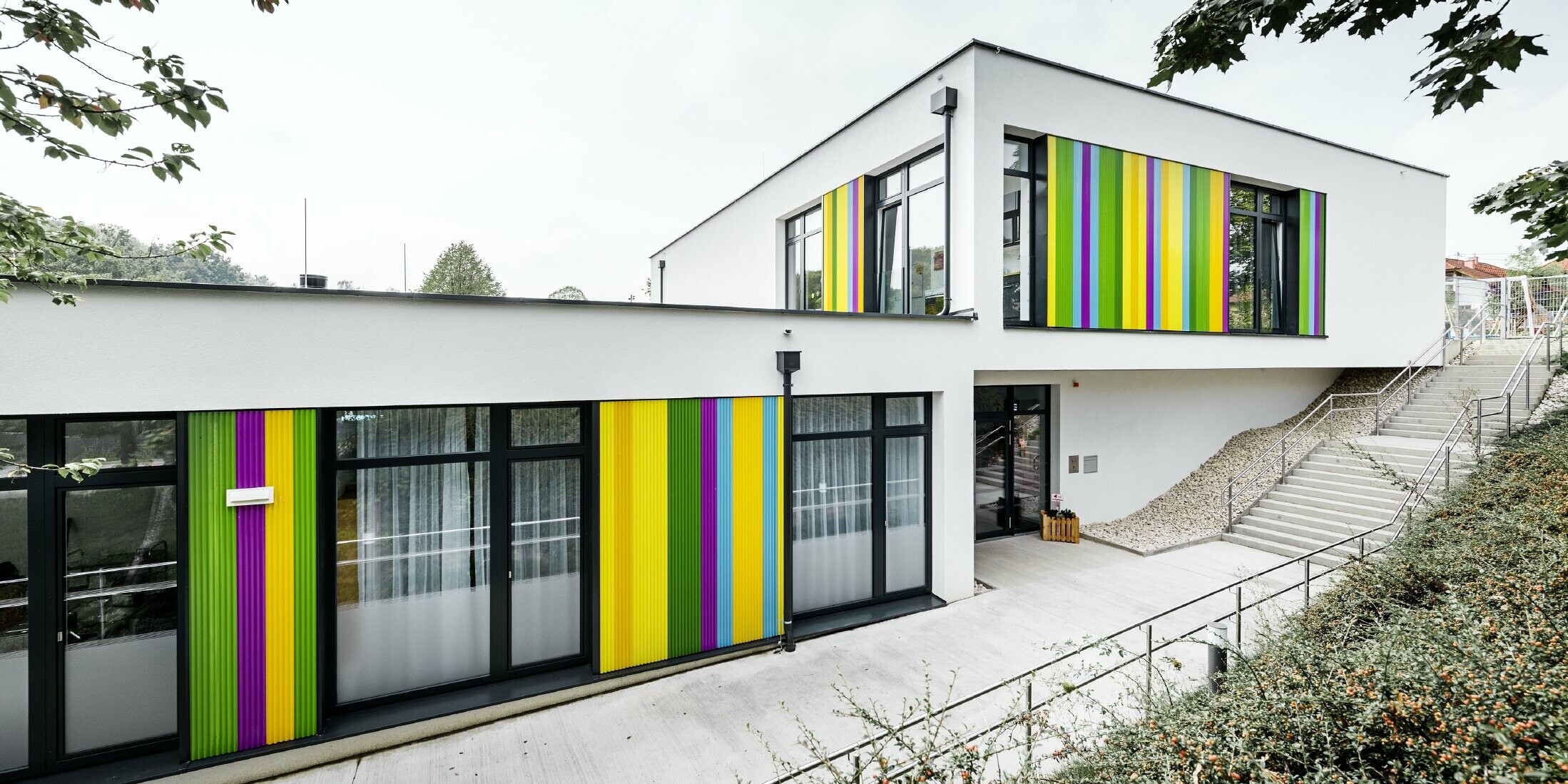 Bunte Fassadenelemente von PREFA für den Kindergarten in Hargelsberg. Das Gebäude hat ein Flachdach und große, bodentiefe Fenster.