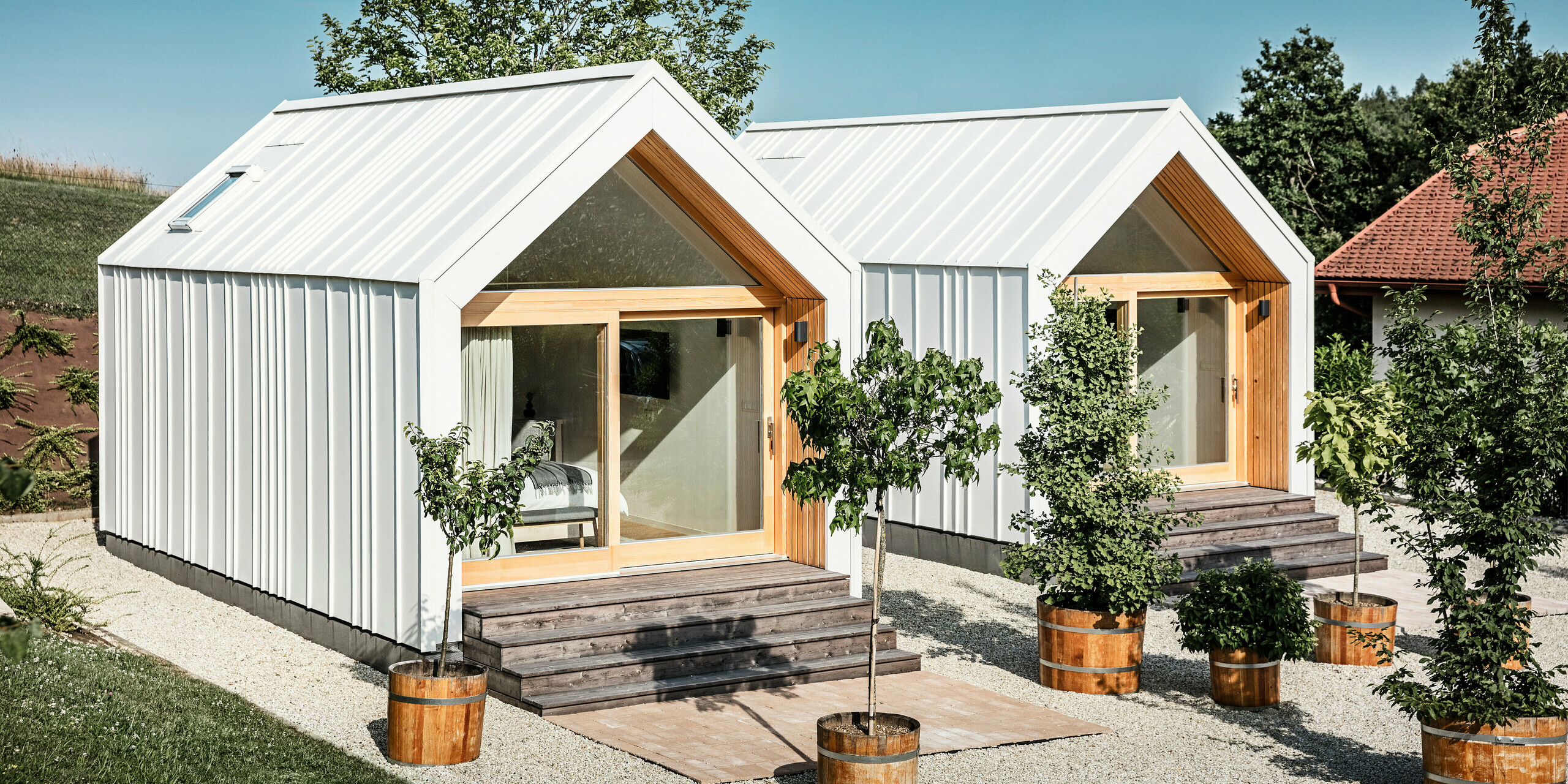 Frontansicht der beiden Tiny Houses der Ferienanlage 'Pri Momi' in Kančevci, Slowenien. Die exklusive Gästeunterkunft wurde mit einer robusten PREFALZ Dach- und Fassadenverkleidung in der Farbe P.10 Reinweiß eingedeckt. Die modernen Mikrohäuser kombinieren eine ästhetische und langlebige Aluminiumhülle mit großen Glasfronten und naturbelassenem Holz und schaffen so eine einladende und warme Atmosphäre. Die Umgebung ist geprägt von gepflegten Rasenflächen und kleinen Bäumen in Pflanzkübeln, die das minimalistische Design der Häuser ergänzen.