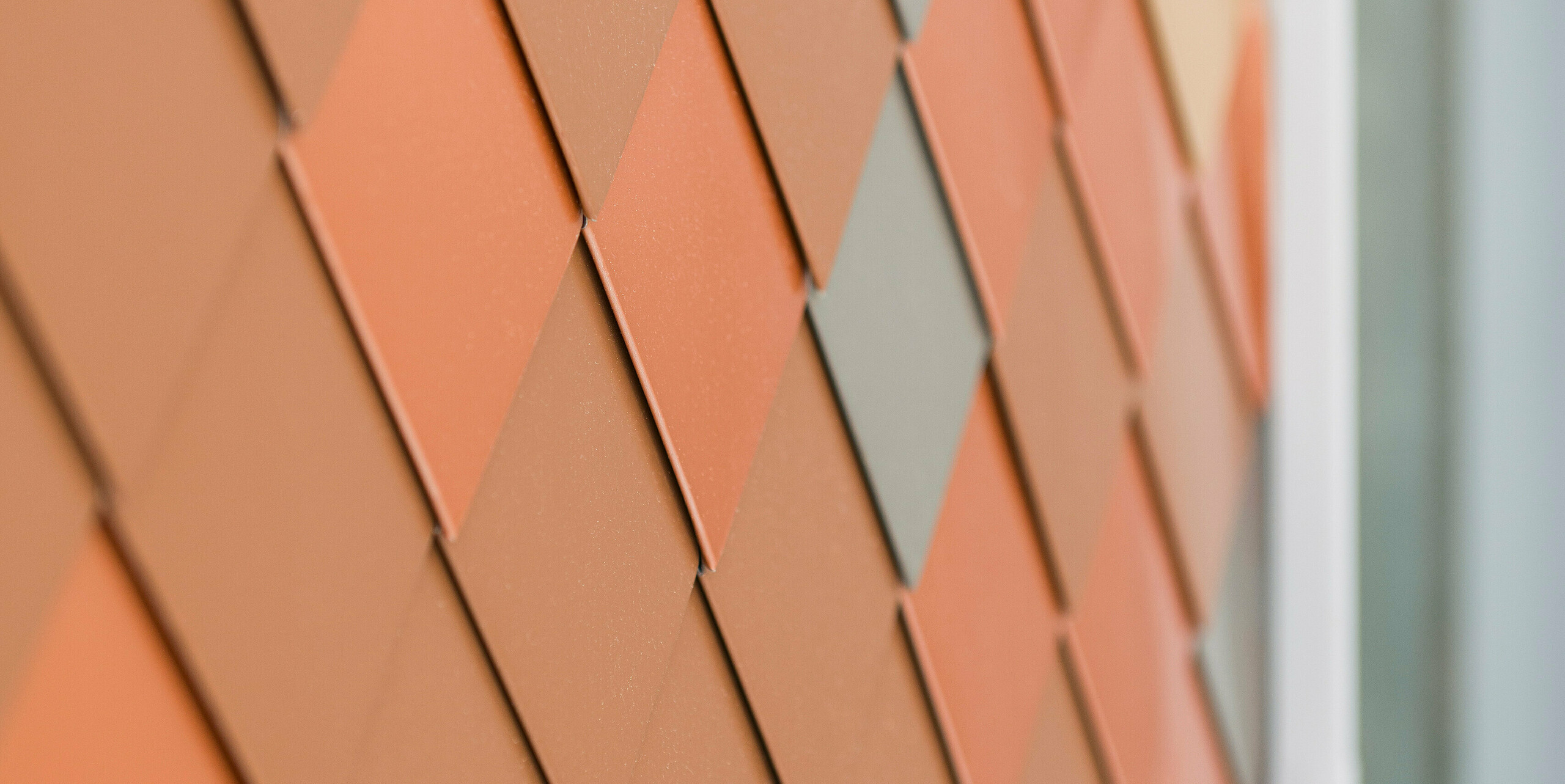 Vue détaillée d'une façade moderne en aluminium PREFA avec un motif géométrique de différentes couleurs. Les petites pièces en aluminium en FALZONAL sont disposées en forme de losange et créent un aspect tridimensionnel qui met en valeur la structure et la profondeur des couleurs.
