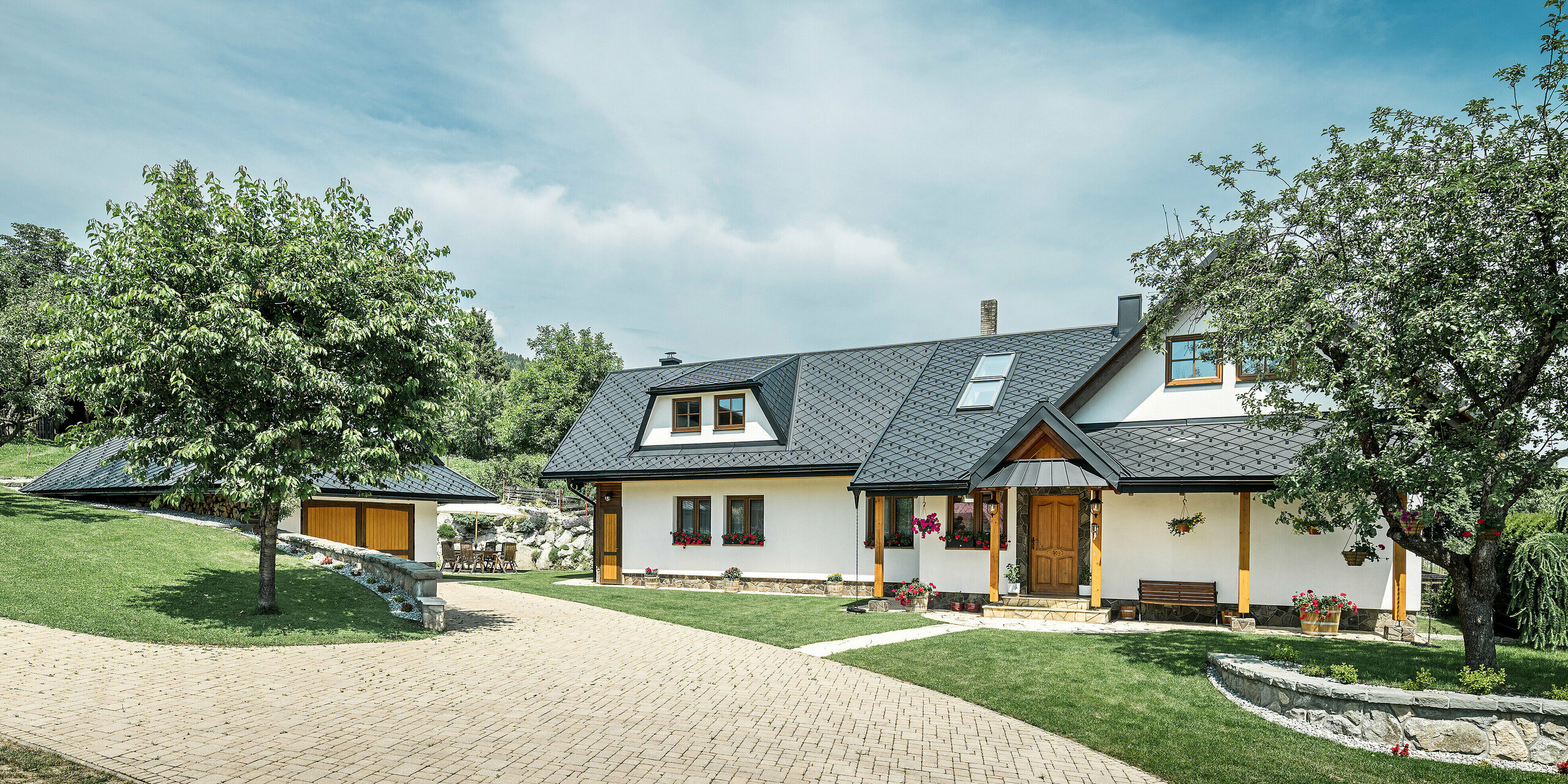 Allée et entrée d'une maison familiale traditionnelle à Stara Bystrica, Slovaquie. Le bâtiment est recouvert de losanges PREFA 29 × 29 en P.10 noir. Les losanges en aluminium assurent une protection robuste et durable. L'allée pavée bien entretenue et le paysage verdoyant environnant, avec un arbre au premier plan, complètent l'aspect général harmonieux de la maison.