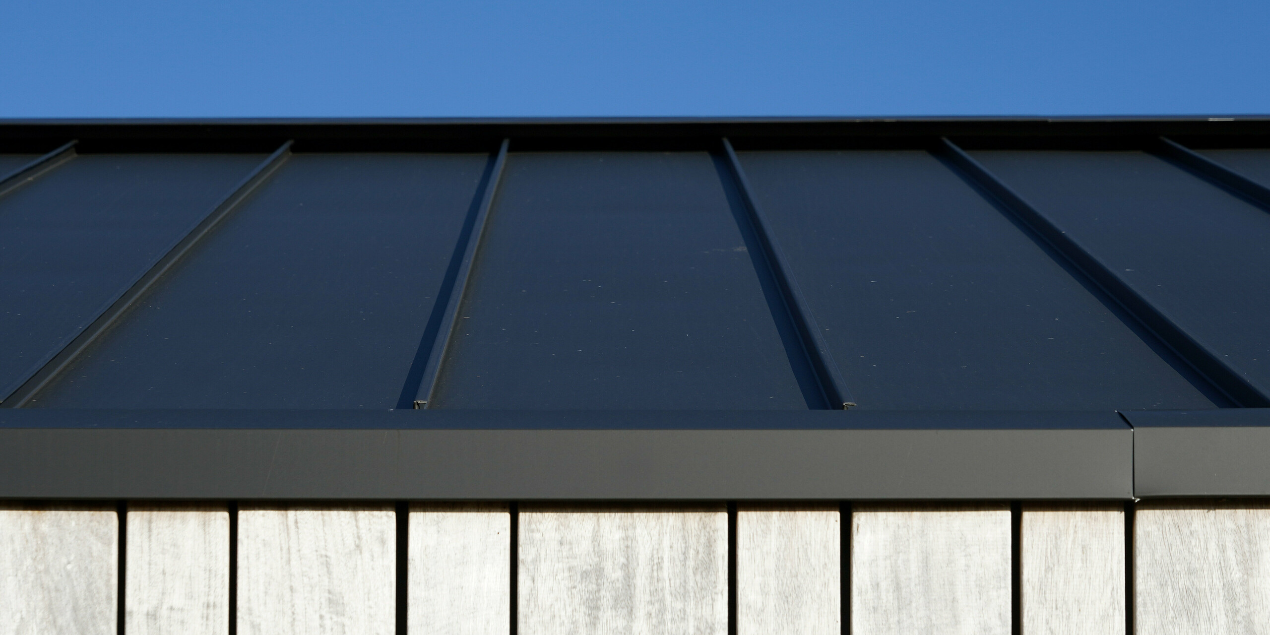 Vue détaillée d'une maison familiale moderne à Uithoorn, aux Pays-Bas, avec toiture à joints debout PREFALZ en gris noir. L'image montre le traitement précis de l'aluminium de haute qualité de PREFA, combinée à des éléments en bois naturel sur la façade. Les lignes claires du toit robuste en aluminium soulignent la construction durable et résistante aux intempéries.