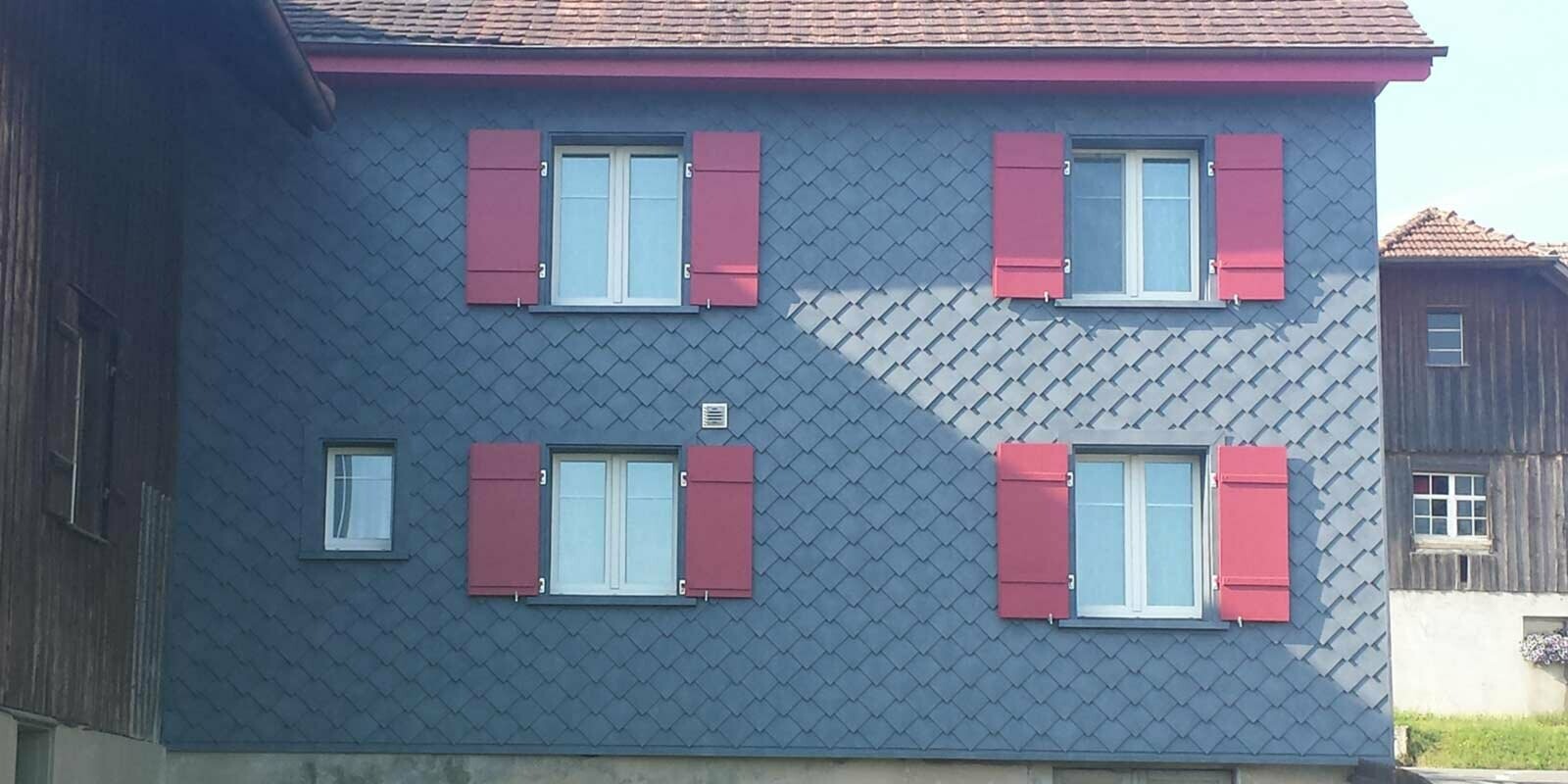 Rénovation de façade à l’aide de losanges de façade PREFA, avec quatre fenêtres dotées de volets rouges