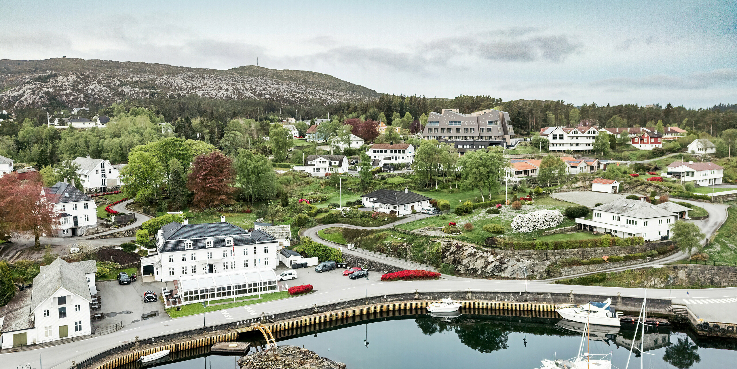 [Translate to Dutch:] Luftaufnahme des Hafens von Bekkjarvik, Norwegen, mit Blick auf das Beckerwyc House im Hintergrund. Das Hotel hebt sich durch seine futuristische Architektur und die Fassade aus PREFA Wandrauten 44 × 44 in Nussbraun von den umliegenden traditionellen weißen Holzhäusern ab. Die malerische Landschaft, der Hafen mit den Booten und die grüne Umgebung unterstreichen die einzigartige Lage des Hotels in dieser idyllischen norwegischen Küstenstadt auf der Insel Selbjørn.