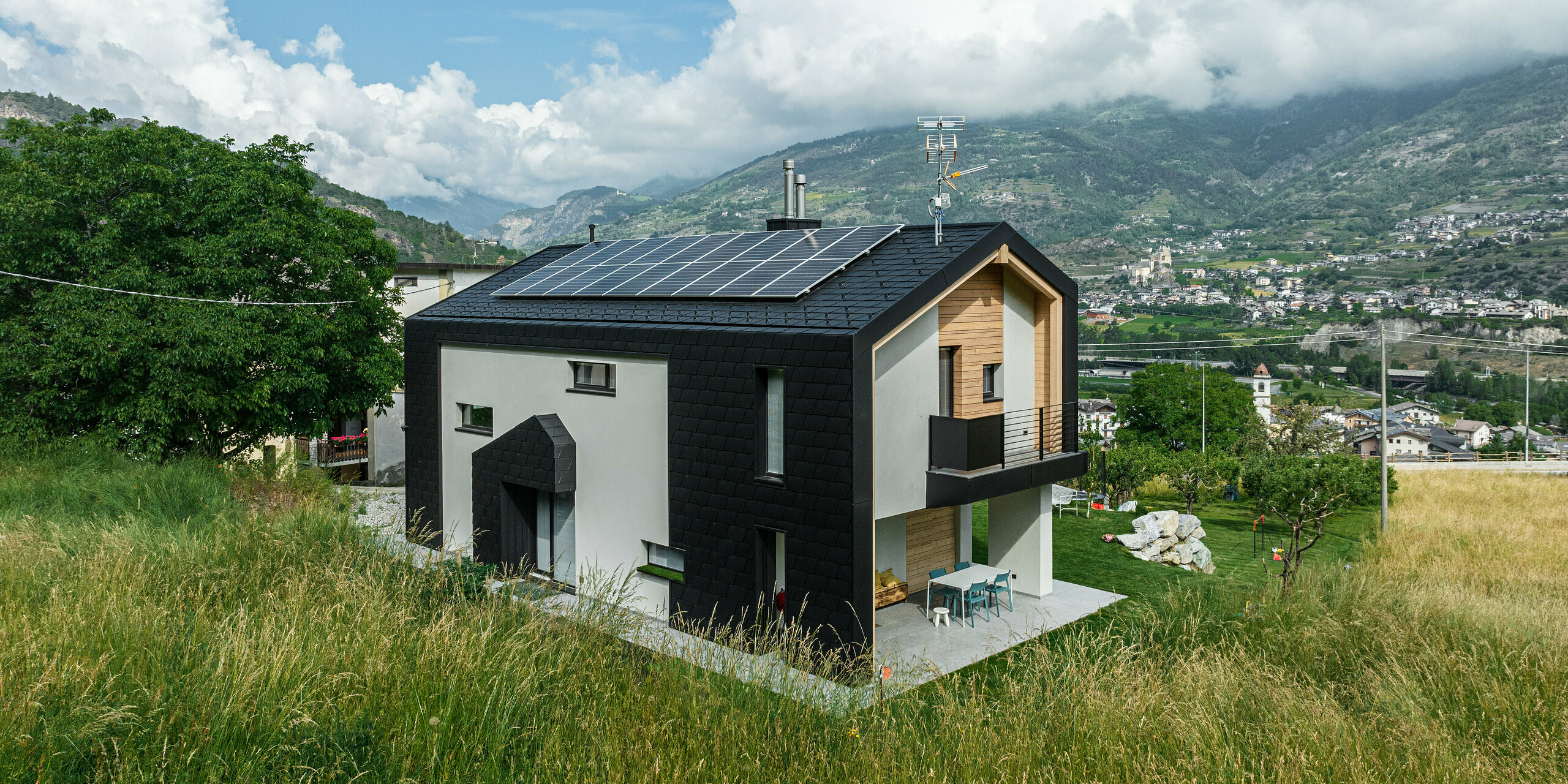 Energieeffizientes Einfamilienhaus mit PREFA Dachschindeln DS.19 und Wandschindeln in P.10 Schwarz. Das moderne Gebäude mit großflächigen Solarpaneelen auf dem Dach ist eingebettet in eine naturnahe Umgebung mit hohem Gras und einem Blick auf das Bergpanorama im Hintergrund. Die Integration von erneuerbaren Energien und langlebigen Baumaterialien unterstreicht das Engagement für Nachhaltigkeit und modernes Wohnen.