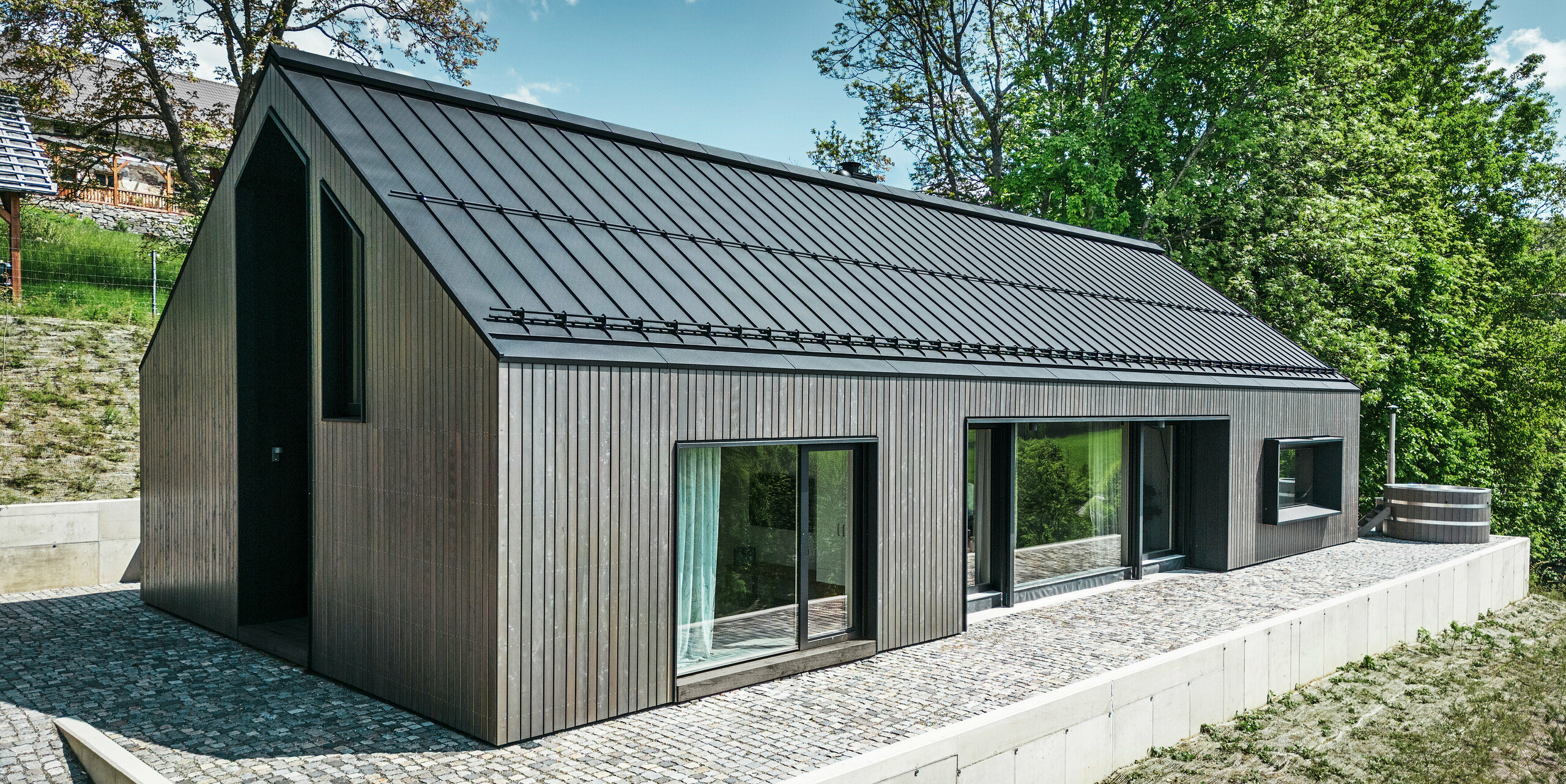 Vorderseite eines Holzmassivhauses in Kuncice, CZ. Das langlebige Aluminiumdach besteht aus PREFALZ in elegantem Schwarz. Das Haus verfügt über großzügige Fensterflächen und einen Whirlpool im Außenbereich.