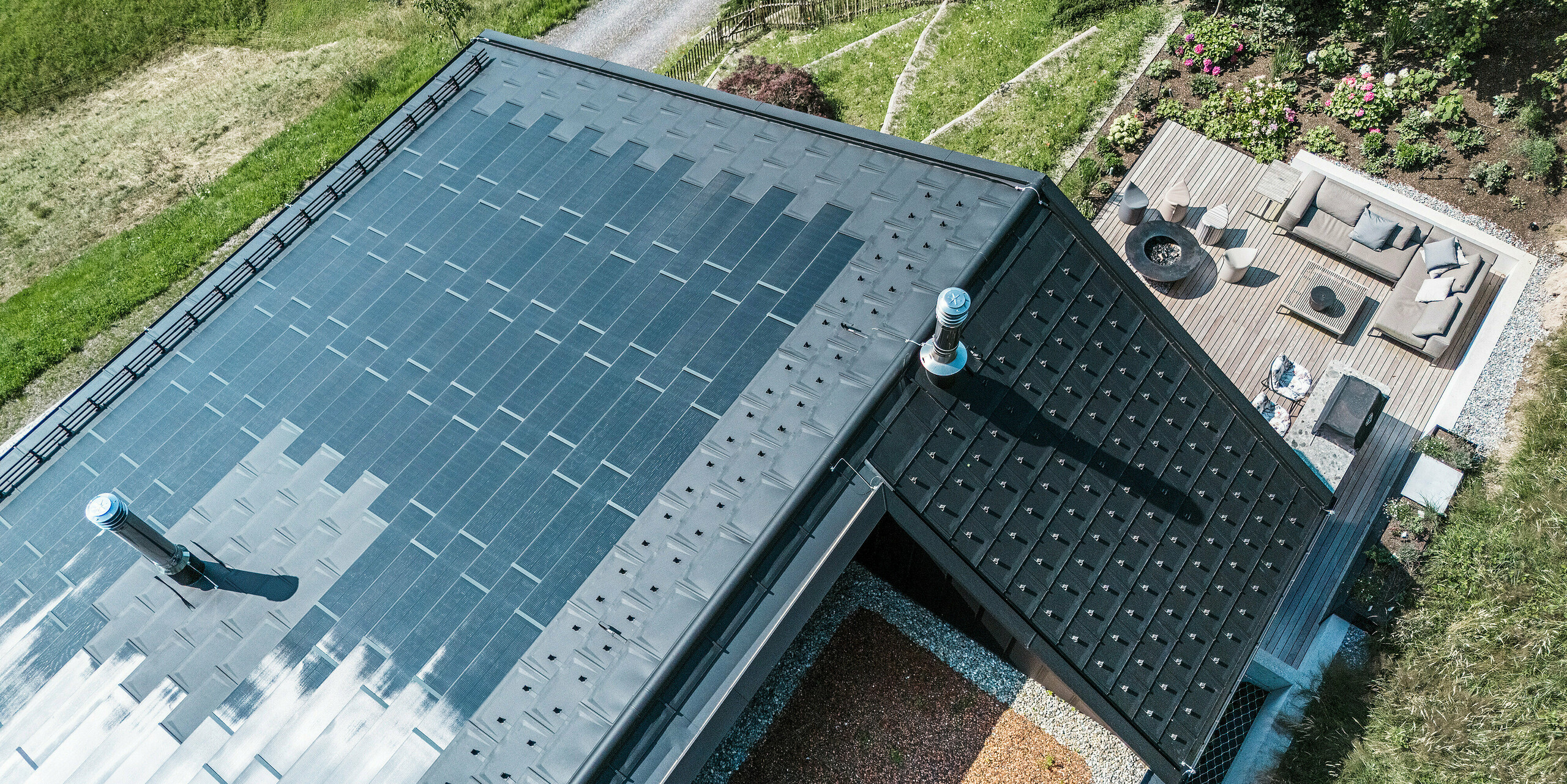 Vogelperspektive auf ein Einfamilienhaus in Egg, Vorarlberg, mit innovativen PREFA Solardachplatten in P.10 Schwarz. Das hochwertige Dachsystem aus Aluminium verfügt über integrierte Photoltaik-Module und kann dadurch Strom aus erneuerbarer Energie erzeugen ohne die homogene Dachlandschaft zu stören. Ergänzt wird das formschöne Satteldach durch robuste PREFA Dachplatten R.16.  Der Schneeschutz sowie die effektive Dachentwässerung mit PREFA Hängerinne und Ablaufrohr garantieren Funktionalität und Langlebigkeit. Am unteren Bildrand ist ein Teil der Dachloggia zu sehen. Am oberen Bildrand auf der rechten Seite ist ein Teil der schönen Terrasse mit hochwertigen Gartenmöbeln zu sehen.