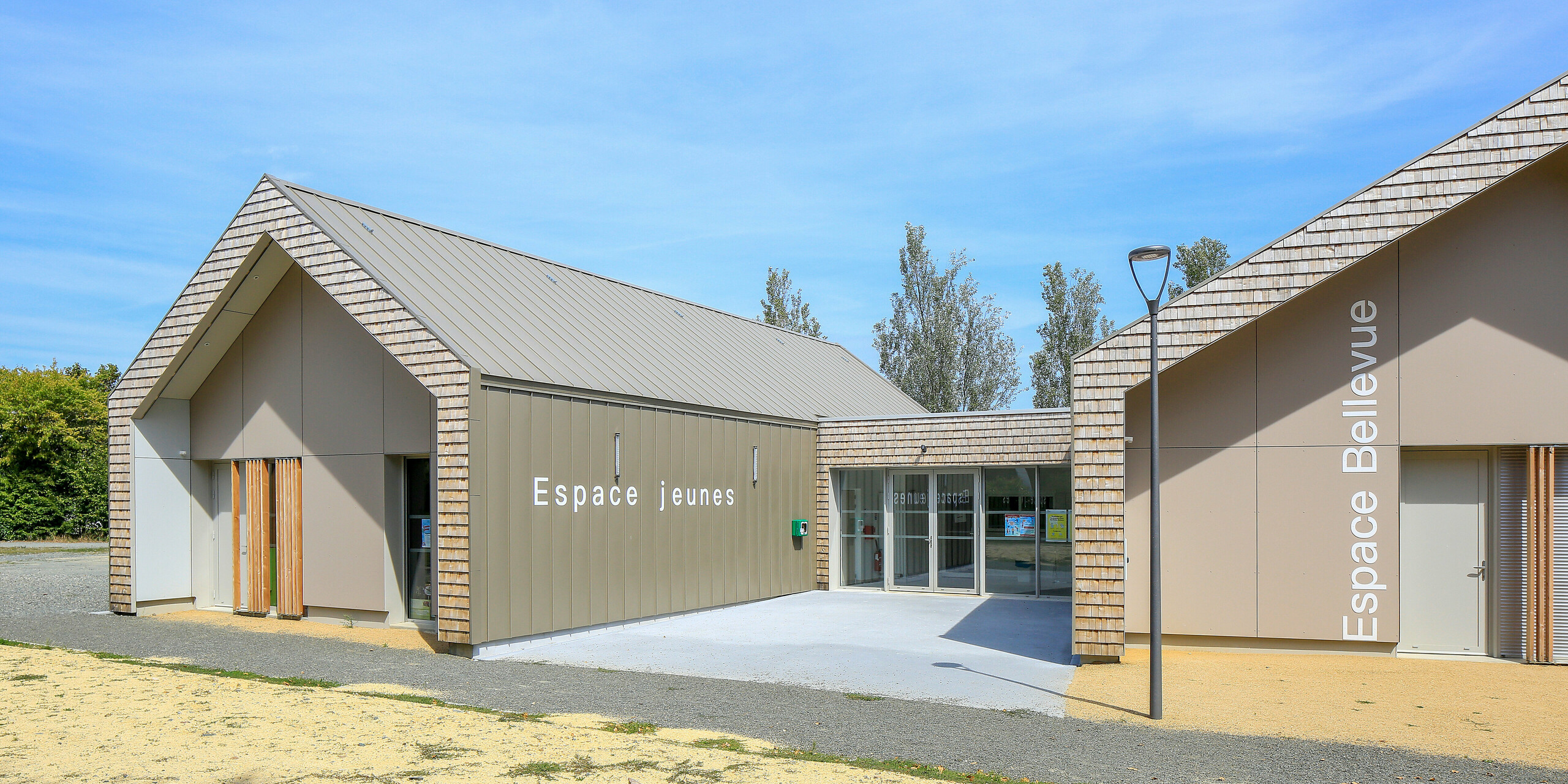 Salles Espace Bellevue à Mûrs-Erigné, France, avec les panneaux 'Espace Jeunes' et 'Espace Bellevue' devant la salle d'événements. Le système de toiture et de façade PREFALZ en bronze décore de grandes parties du centre de jeunesse moderne. Le bardage à joint debout en aluminium résistant aux intempéries de PREFA offre une solution durable et moderne pour les projets de construction. La combinaison du revêtement en aluminium et de l'aspect bois naturel s'intègre harmonieusement dans l'environnement.