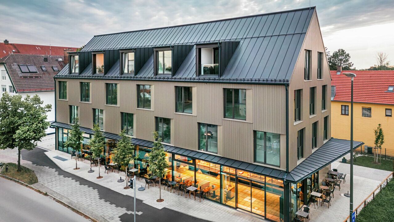 La photo montre un bâtiment moderne avec un revêtement de façade en Prefalz de couleur bronze et un toit à deux versants, également recouvert de Prefalz de couleur gris noir. La grande façade vitrée du rez-de-chaussée est encadrée d'arbres et crée une atmosphère invitante pour le restaurant situé en contrebas. La combinaison des matériaux Prefalz de haute qualité et de l'architecture moderne confère au bâtiment un aspect élégant et contemporain.