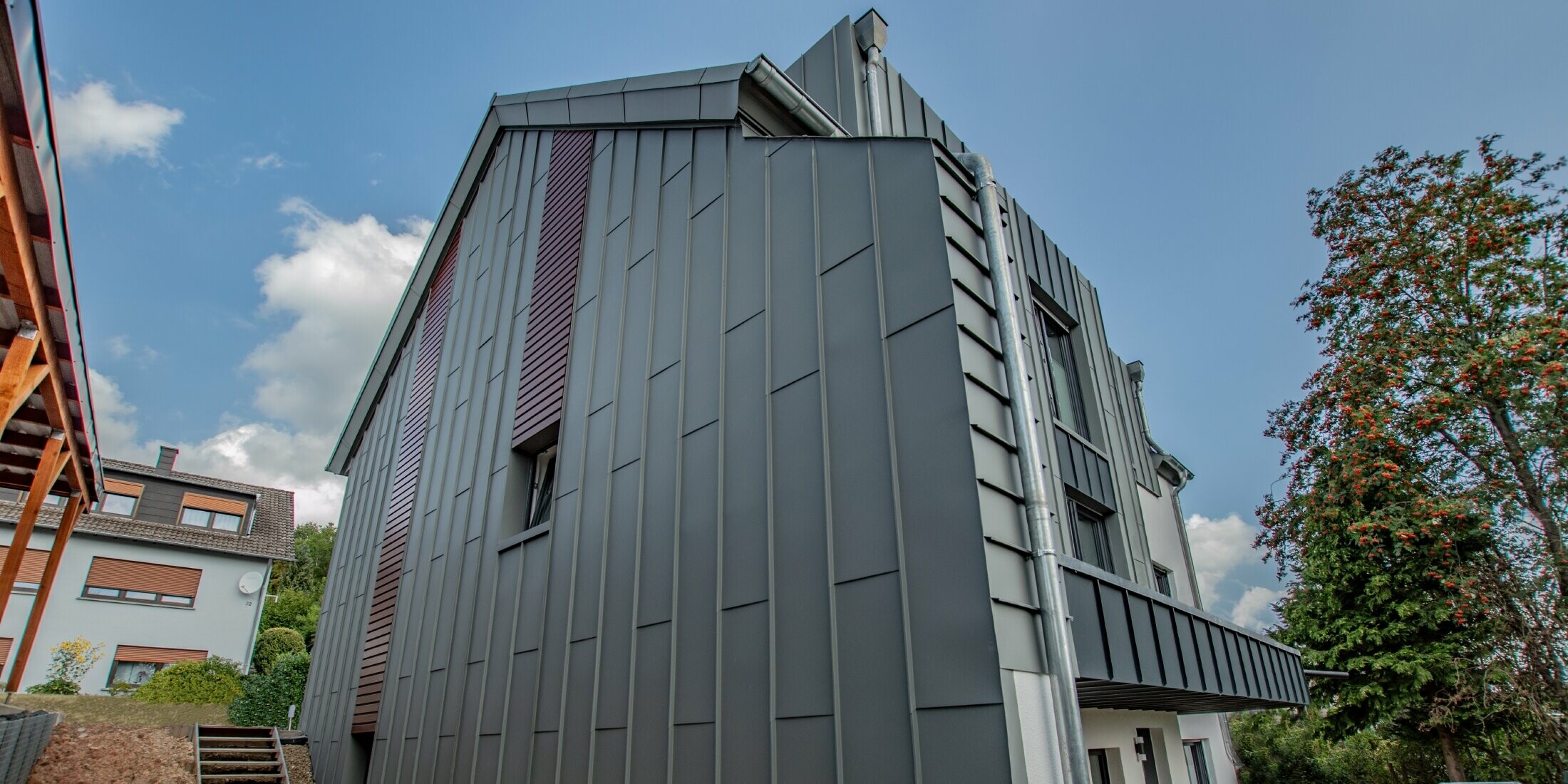 Fassadengestaltung mit vertikaler Winkelstehfalzfassade in hellgrau und Fassadenpaneelen, horizontal verlegt, in Holz dunkel. Die gesamte Fassade wurde aus Aluminium von PREFA gefertigt.