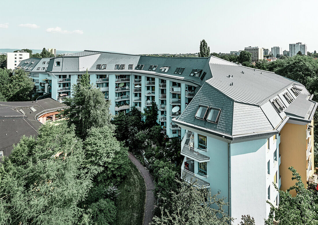 Grand bâtiment d’habitation consacré à l’accueil des personnes âgés, recouvert de bardeaux de toiture et Prefalz PREFA couleur gris pierre, avec de nombreux balcons.