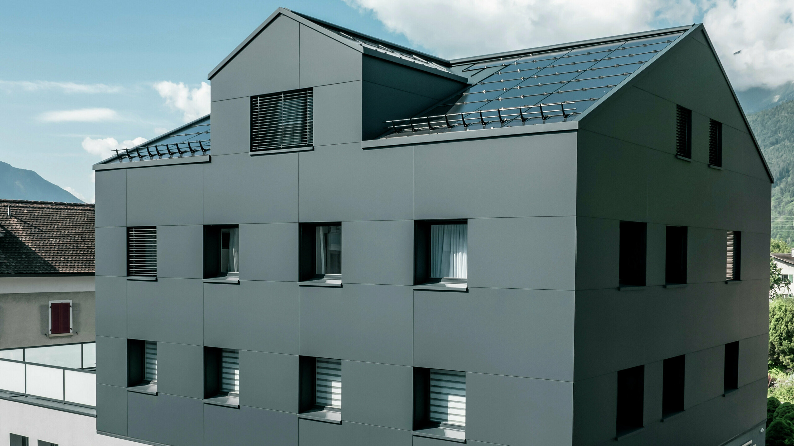 Modernisiertes Wohn- und Geschäftsgebäude nach der Fassadensanierung mit PREFABOND Aluminium Verbundplatten in Anthrazit. Die klare, elegante Linienführung der neuen Fassade verleiht dem Gebäude ein modernes Erscheinungsbild. Im Erdgeschoss befindet sich das Architekturbüro Voser. Die Umgebung mit Bäumen und Bergen rundet die harmonische Optik ab.