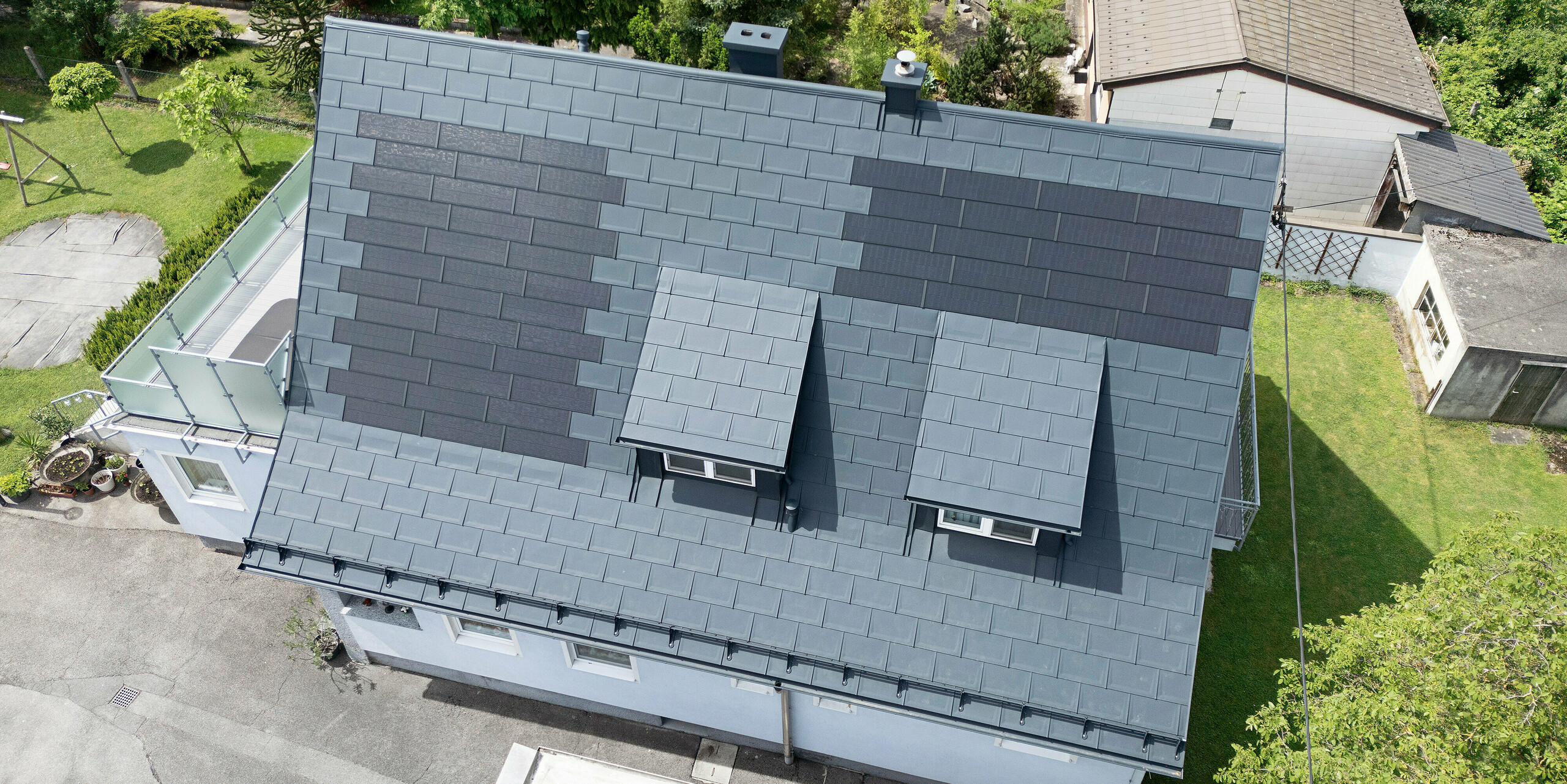 Toiture entièrement rénovée d'une maison familiale à Leonding avec des tuiles solaires PREFA en anthracite. La vue plongeante montre le toit modernisé avec des modules solaires intégrés fabriqués à partir de produits en aluminium PREFA. Cette solution durable et résistante aux intempéries offre un moyen durable et économe en énergie de produire de l’électricité. L'image illustre l'intégration esthétique des modules solaires dans la surface du toit, qui soutient une architecture moderne et fonctionnelle. Les tuiles solaires innovantes ont été placées autour de deux lucarnes.