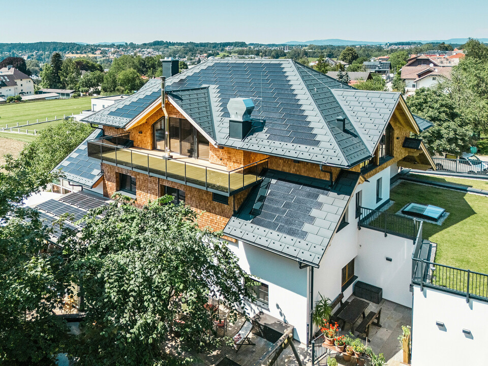 Vue aérienne du Zenz'n Stub'n à Schörfling am Attersee avec un toit récemment rénové en tuiles solaires PREFA et panneaux de toit R.16 en P.10 anthracite. La toiture en tôle de haute qualité allie un design moderne à une production d'énergie durable. Le balcon spacieux et la gouttière harmonieusement intégré soulignent l'aspect élégant du bâtiment.