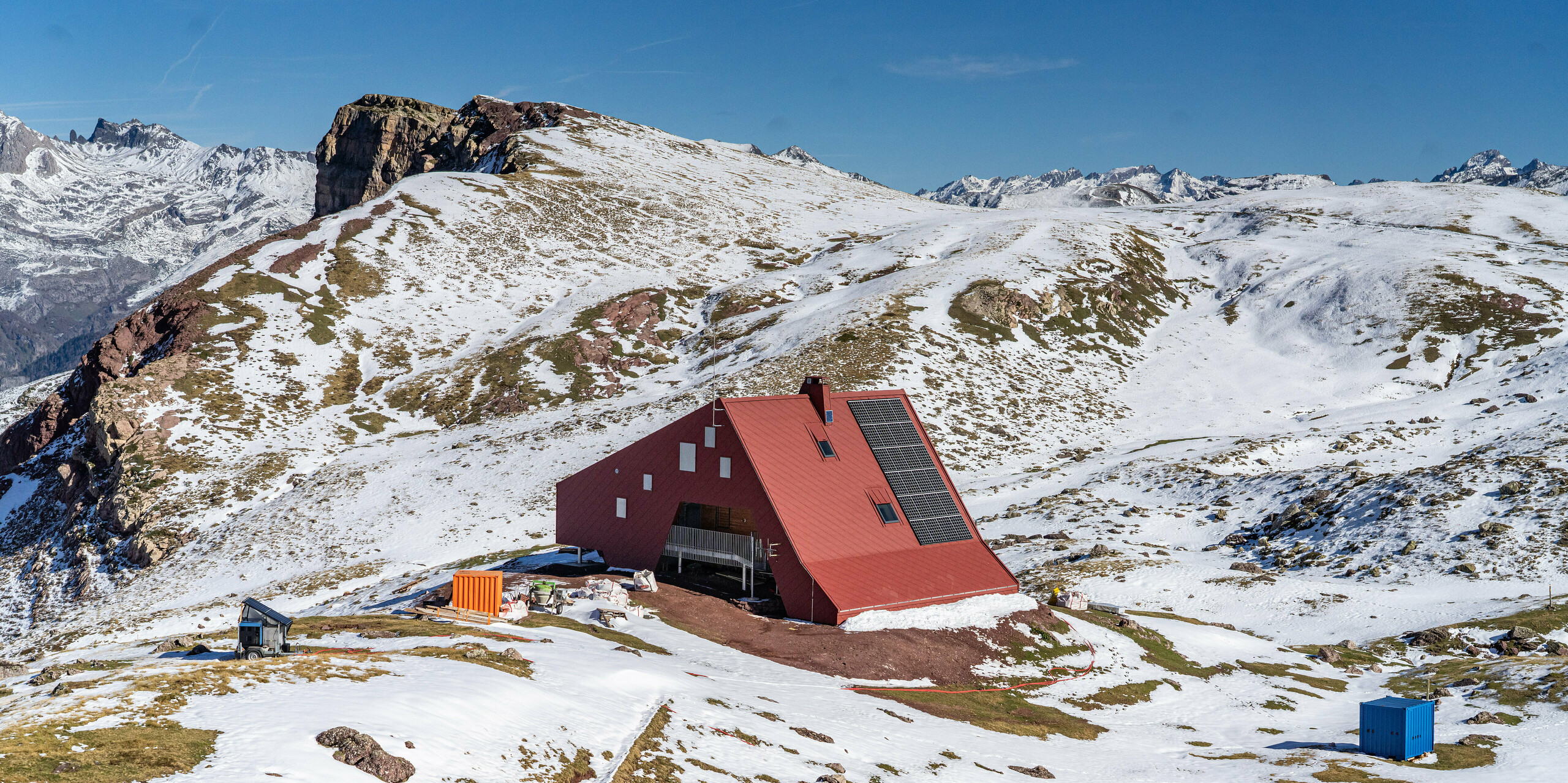 Vue plongeante sur le refuge d'Arlet dans le Parc National des Pyrénées avec un paysage de montagne enneigé autour du bâtiment rouge oxyde avec un toit et une façade en aluminium