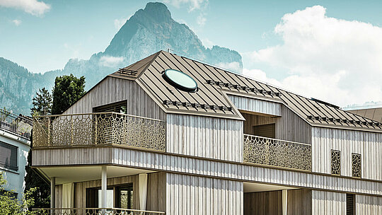 Vue détaillée d'un immeuble d'habitation moderne à Liebwylen, Suisse, avec un toit en aluminium PREFALZ de couleur brune et une fenêtre de toit ronde. La façade d'aspect bois et les balustrades artistiquement conçues s'harmonisent avec l'imposant décor de montagne en arrière-plan.