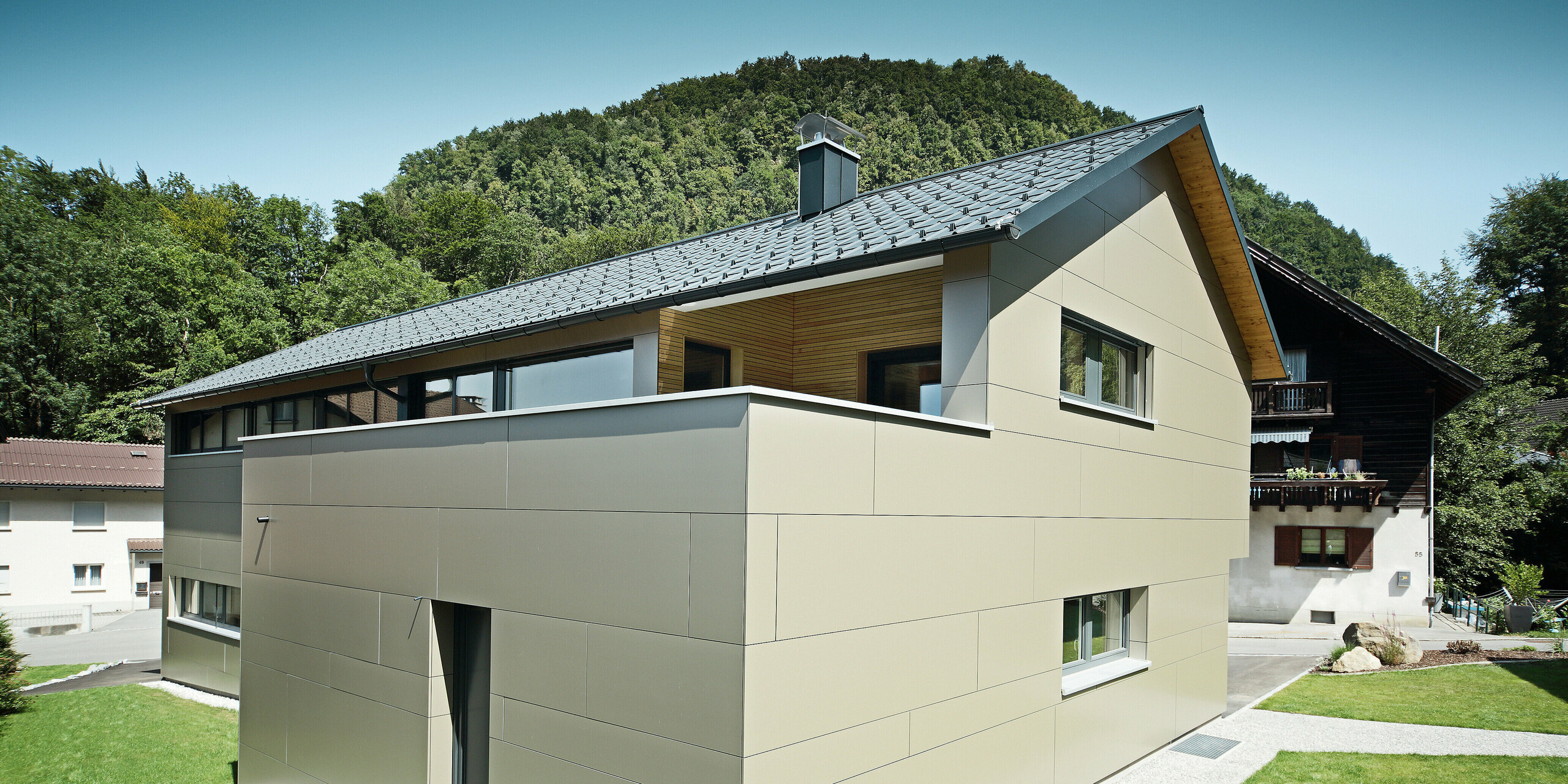 Einfamilienhaus in Hohenems mit einer stilvollen PREFA Fassade aus Aluminium Verbundplatten in Bronzemetallic und einem harmonierenden Dach aus PREFA Dachplatten in P.10 Anthrazit. Das Bild präsentiert das Gebäude in voller Pracht vor einem bewaldeten Hintergrund, wobei die saubere Linienführung und die Farbkombination der Materialien die zeitgenössische Architektur unterstreichen und für einen modernen, langlebigen und wartungsarmen Gebäudeschutz sorgen.