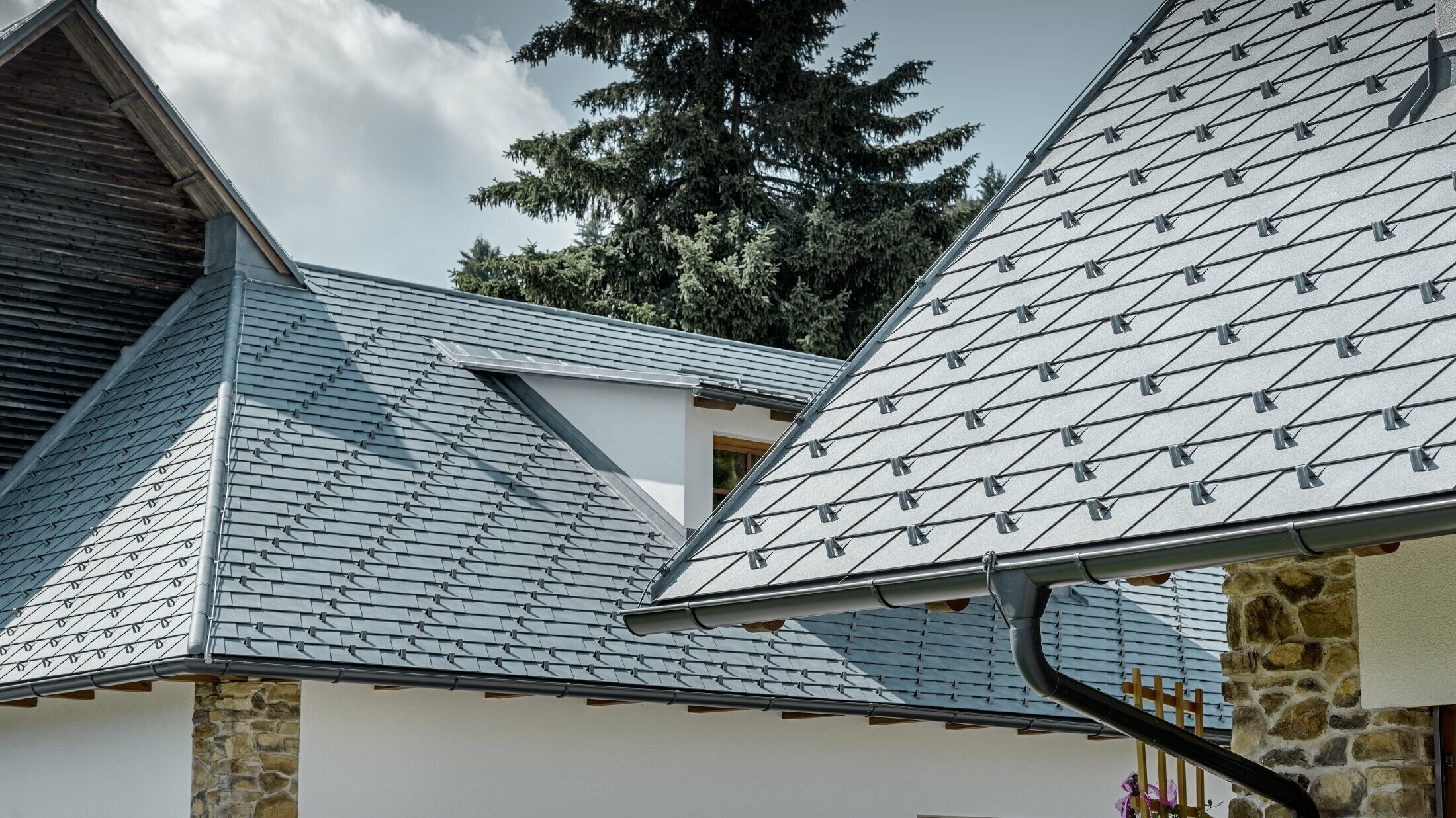 Detailopname van een aluminium dakbedekking van PREFA; dakschindel in steengrijs met de aluminium dakgoot in antraciet van PREFA; op de achtergrond herkent men een dakkapel met lessenaarsdak met staand felsdak. De gevel is wit met ingewerkte steenelementen.