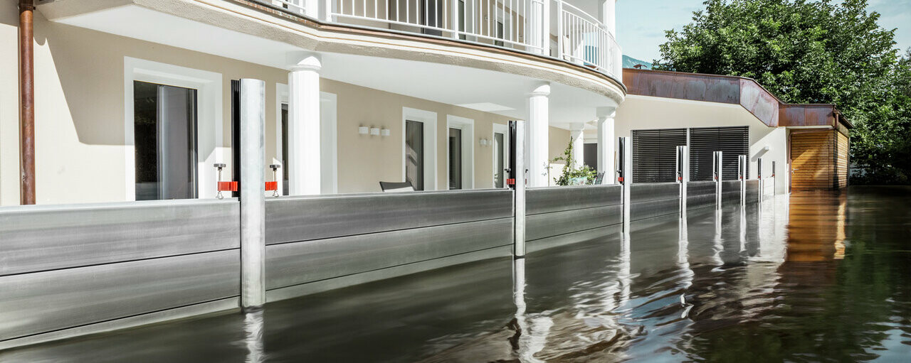 De afbeelding toont een vrijstaand huis met een balkon. Het PREFA hoogwaterbeschermingssysteem beschermt het huis tegen het al stijgende overstromingswater.