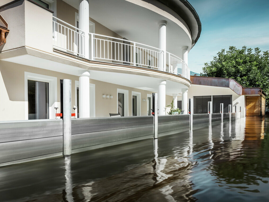 De afbeelding toont een vrijstaand huis met een balkon. Het PREFA hoogwaterbeschermingssysteem beschermt het huis tegen het al stijgende overstromingswater.