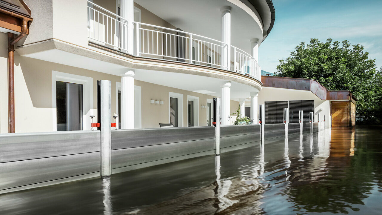 De afbeelding toont een vrijstaand huis met een balkon. Het PREFA hoogwaterbeschermingssysteem beschermt het huis tegen het al stijgende overstromingswater.